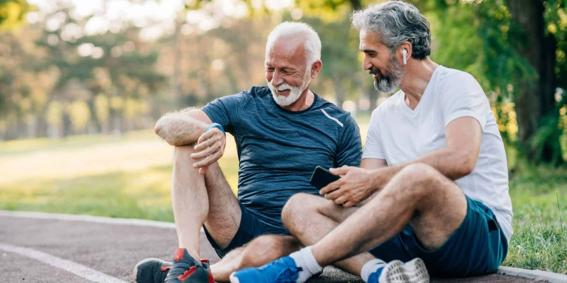 Zwei ältere Freunde, die nach dem Joggen auf einer Laufstrecke sitzen und die Zeit auf einer Smartwatch überprüfen.